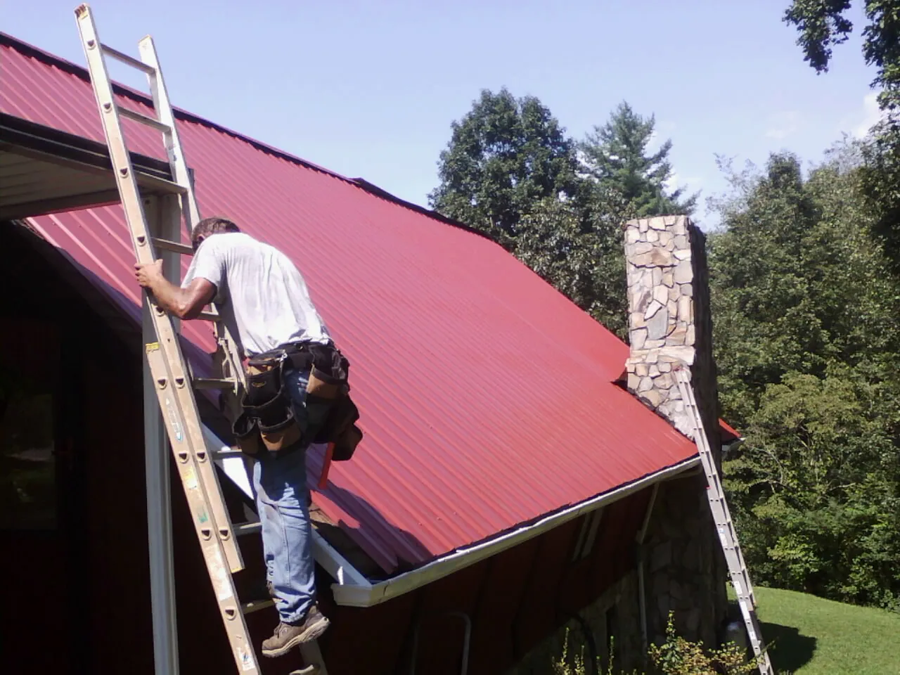 Roofer on a ladder beside a red metal roof and stone chimney — Soffit & Fascia Repair by Roofing Brothers in Rossville